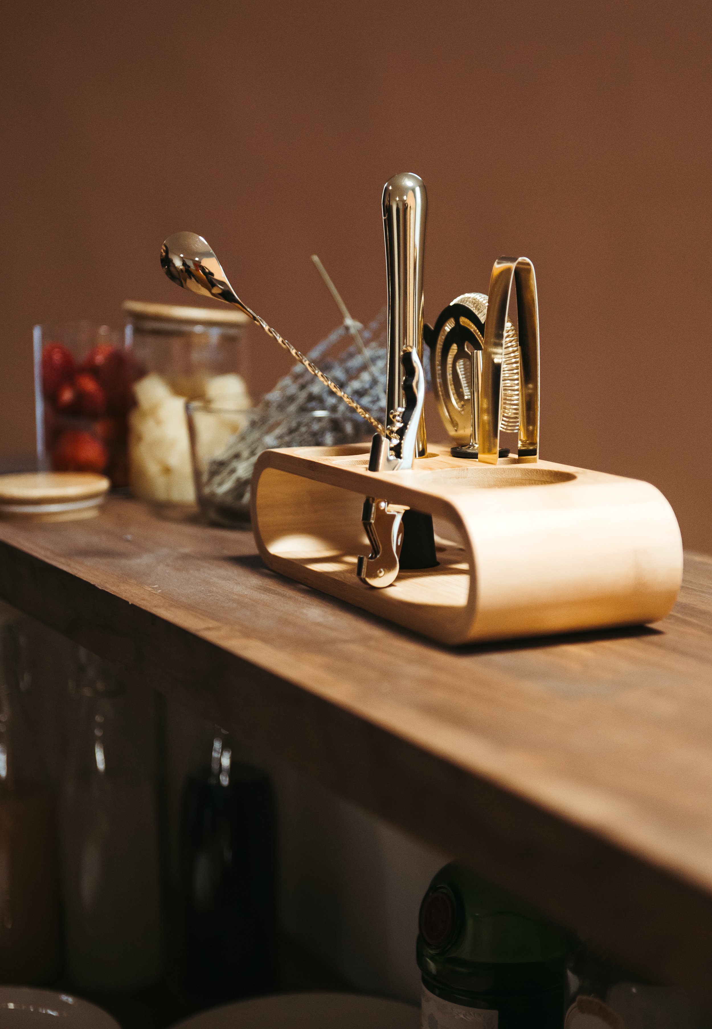 Bar countertop with a wooden bar tool holder containing various tools, a spoon, and a pair of tongs. In the background, there are jars of strawberries and pineapples, a spoon, and some napkins.