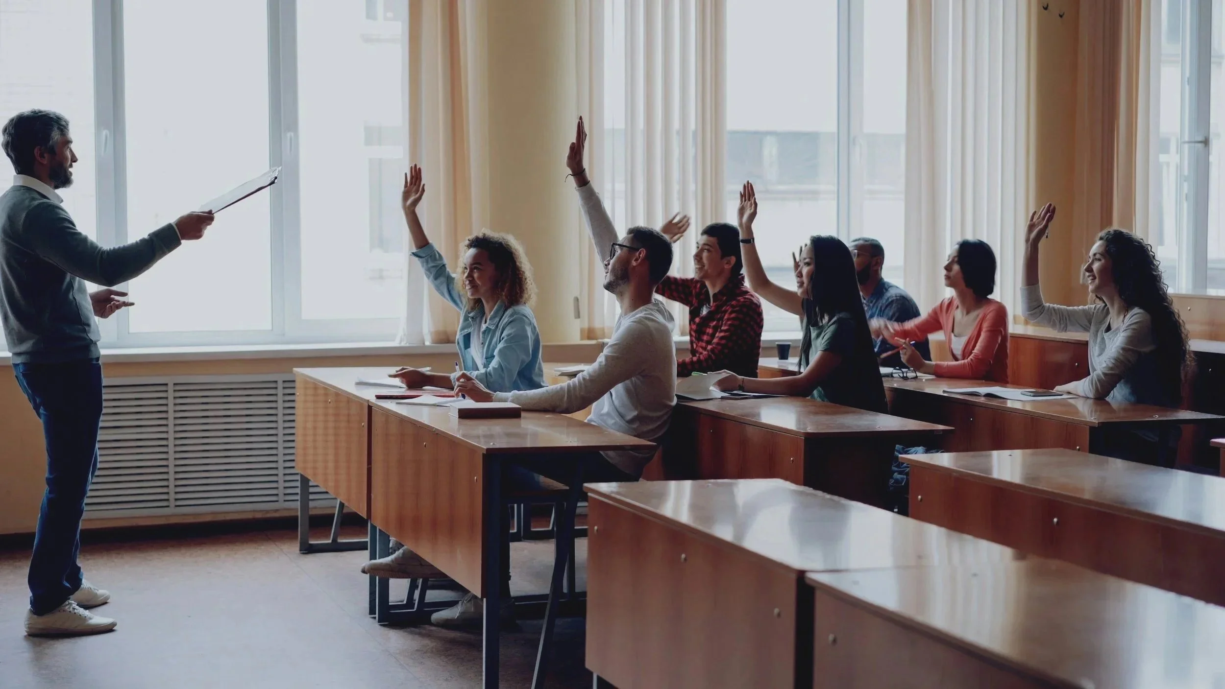 A classroom with students raising their hands while a teacher stands at the front with a clipboard, engaging in a discussion.