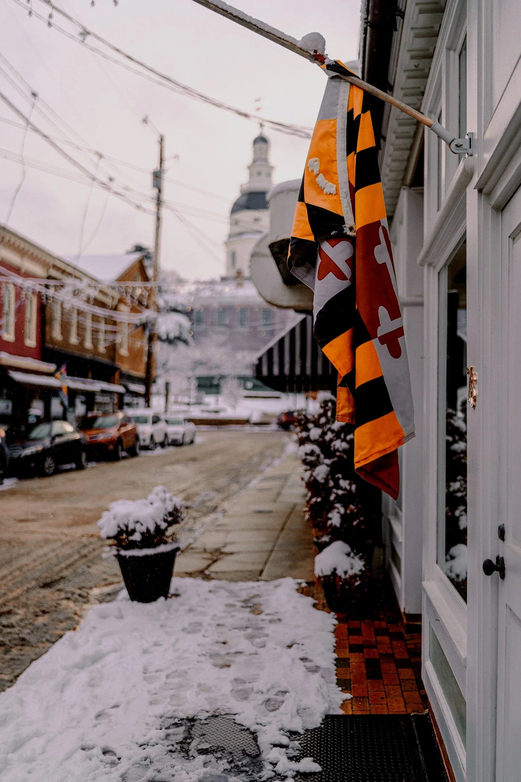 A snow-covered sidewalk outside a building with a black and orange flag hanging on the wall. There are small snow-covered potted plants along the sidewalk and parked cars in the distance. A church steeple is visible in the background.