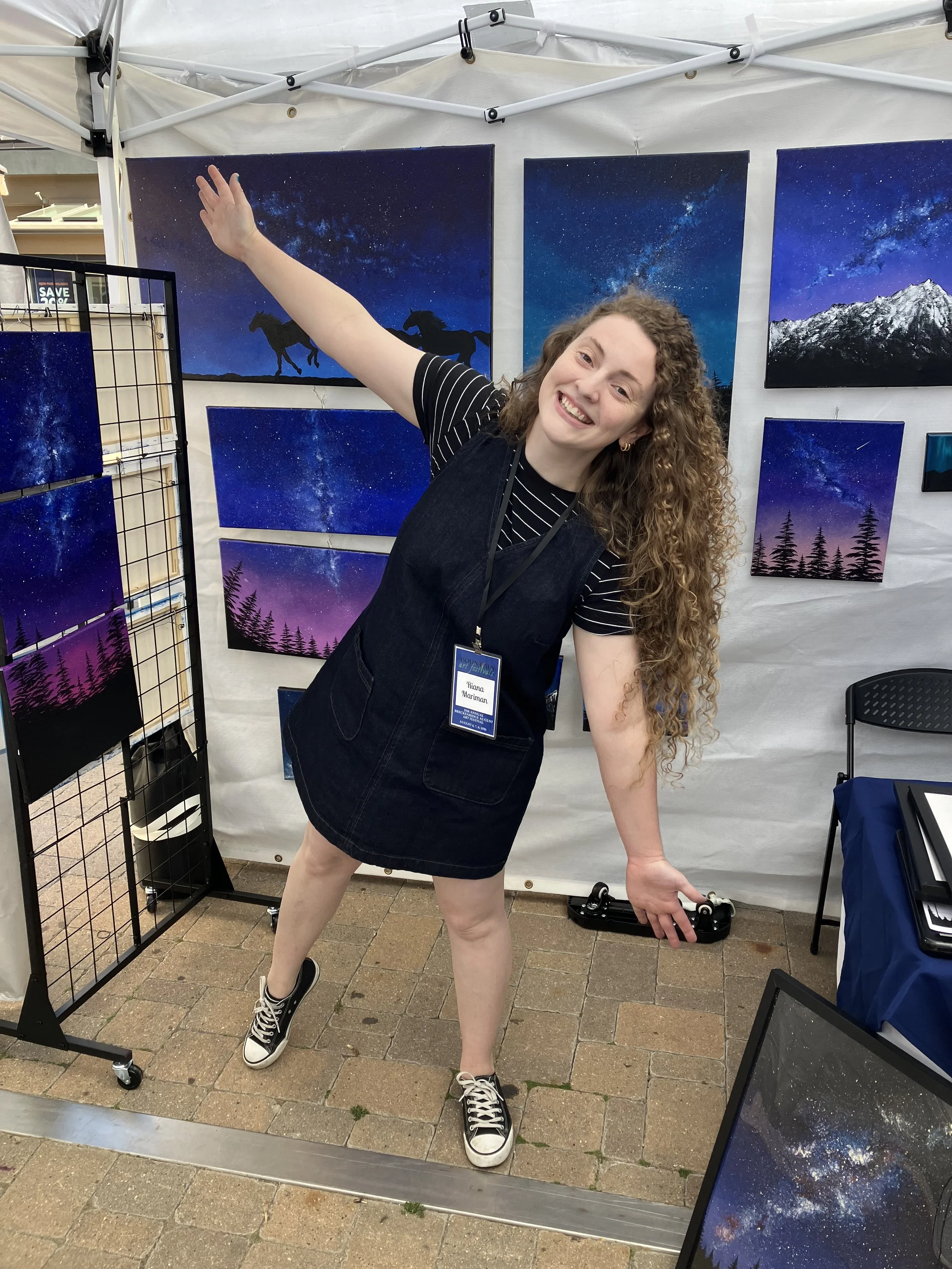 A woman with curly hair smiling and posing at an art fair booth with space-themed paintings of starry skies and mountain landscapes behind her.