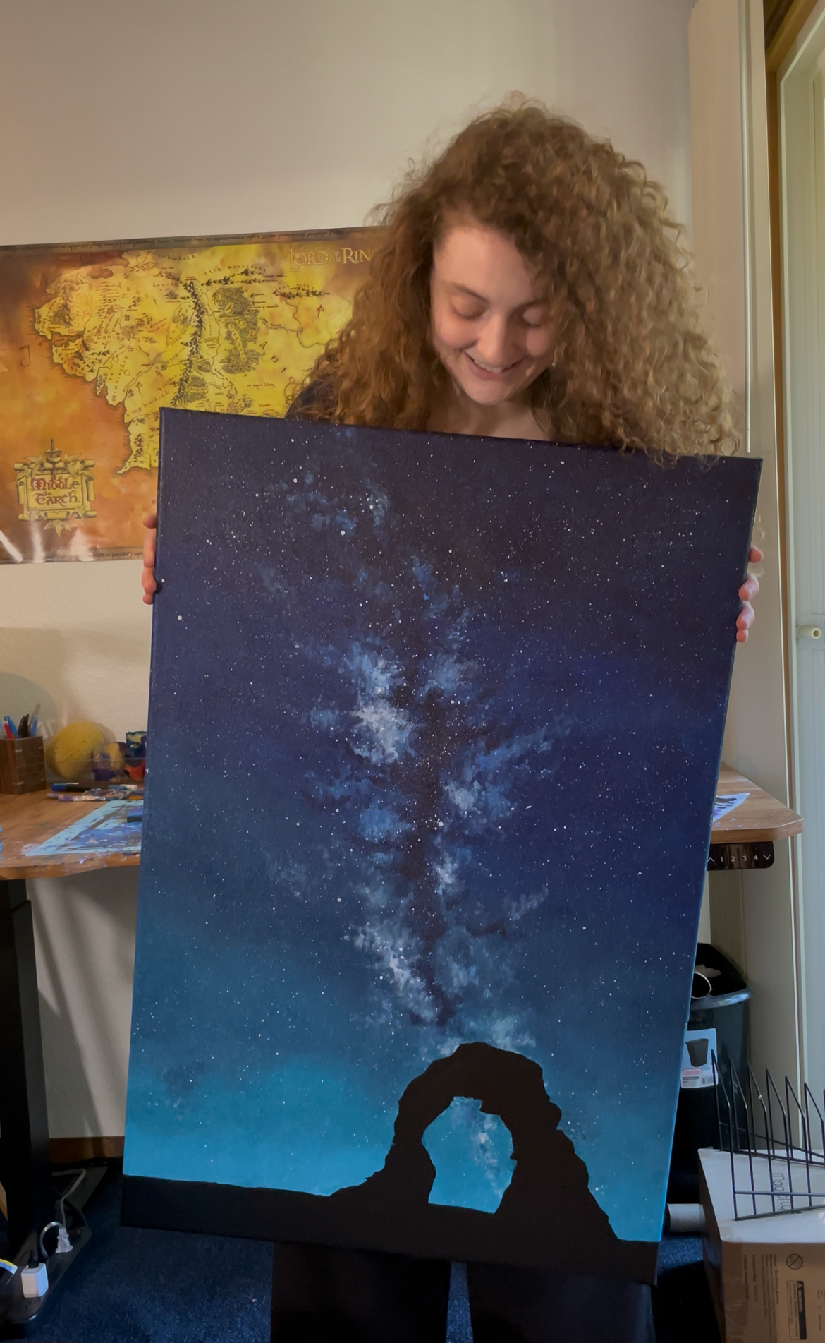 Person with curly hair holding a painting of the Milky Way galaxy at night, with a rock formation at the bottom background.