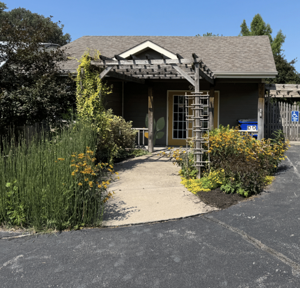 A small dark gray house with a beige door and a yellow frame, surrounded by lush green plants and yellow flowers. A wooden garden archway is over the sidewalk leading to the front door. There is a blue wheelchair accessibility sign and two recycling bins to the right of the house. The sky is clear and blue.