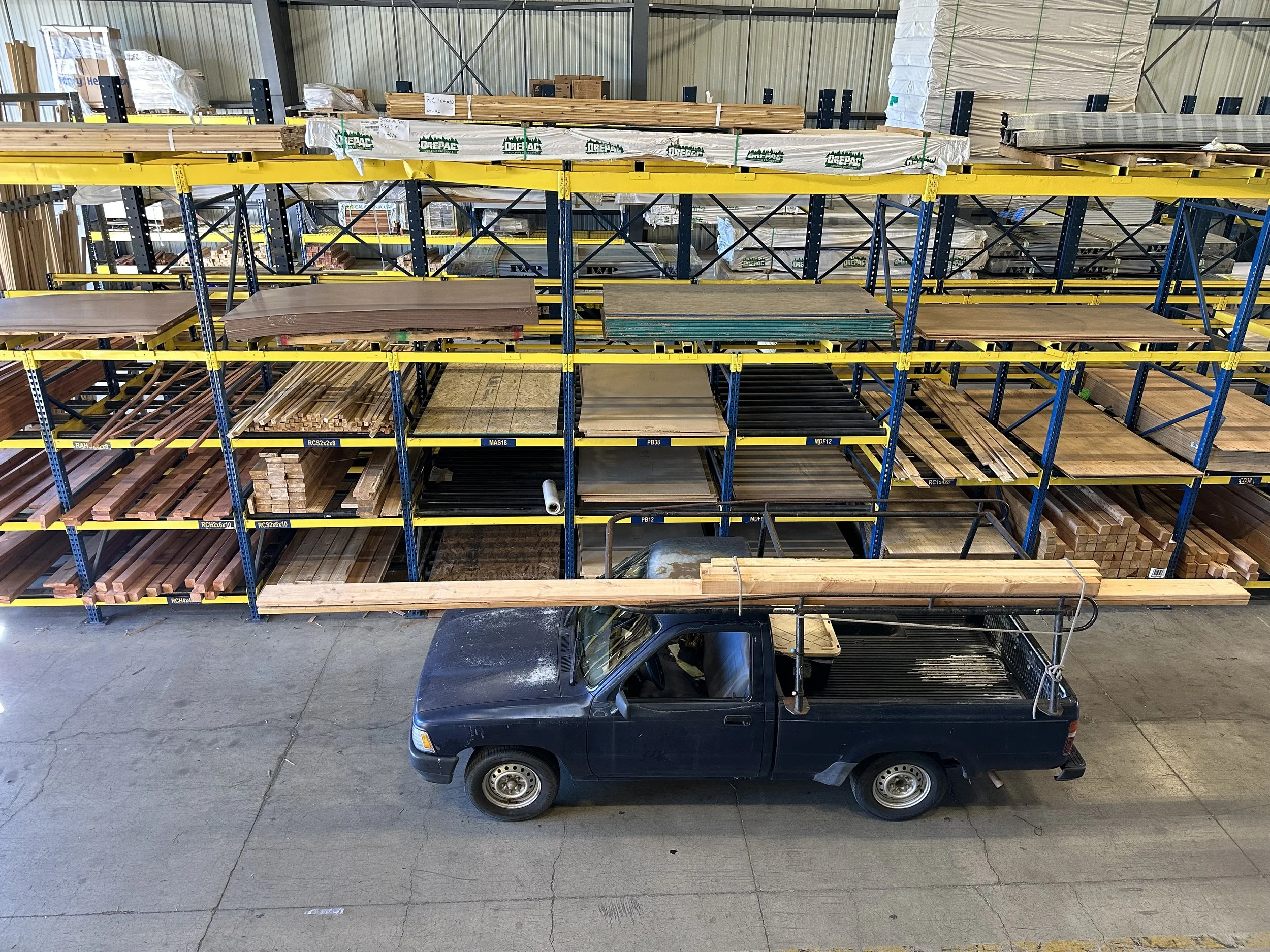 A black pickup truck with an open bed parked underneath metal storage racks filled with various types of wood and building materials in a warehouse.