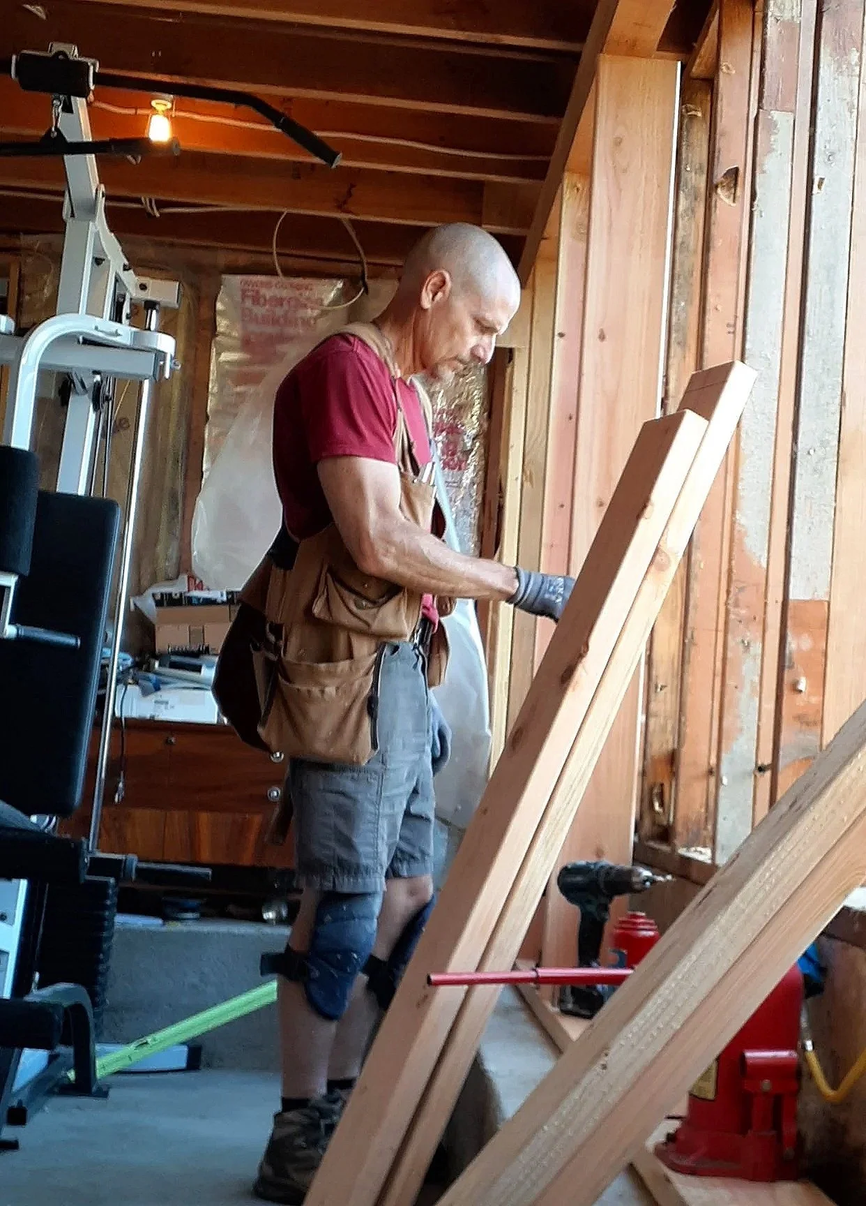 A man wearing a red t-shirt, work apron, jean shorts, and knee pads is working on a carpentry project in a workshop with wooden framing. He is focused on a piece of wood and appears to be measuring or cutting.