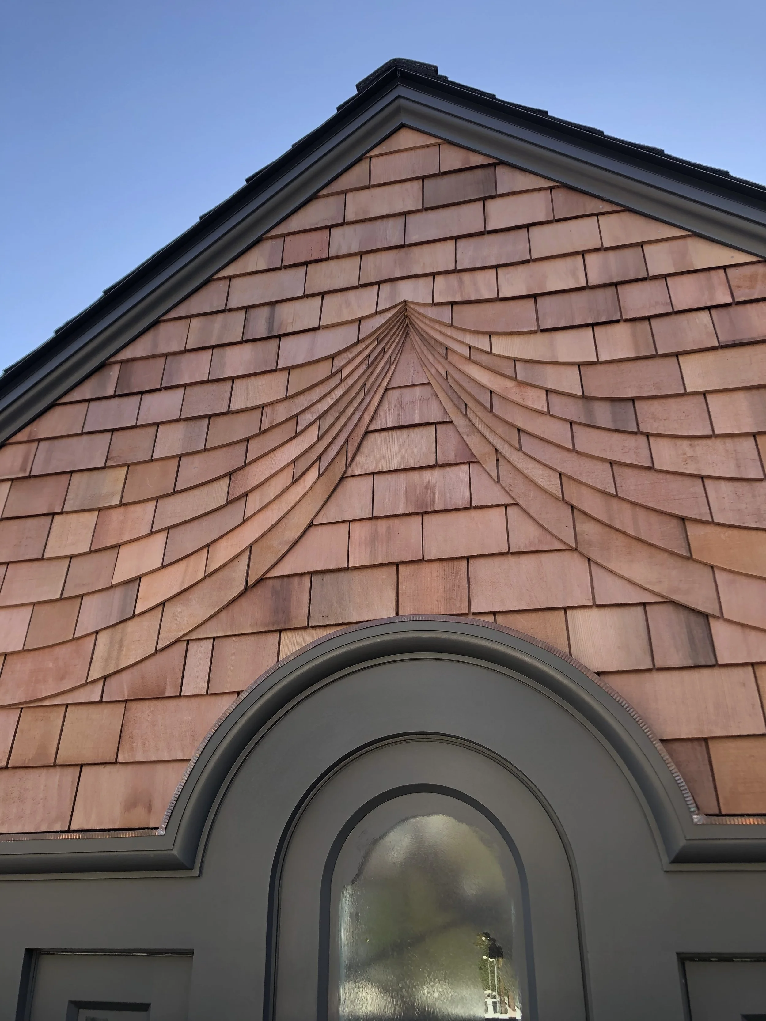 Close-up view of a house with a shingled gable roof and arched window, featuring pinkish-brown shingles and grey trim, against a clear blue sky.
