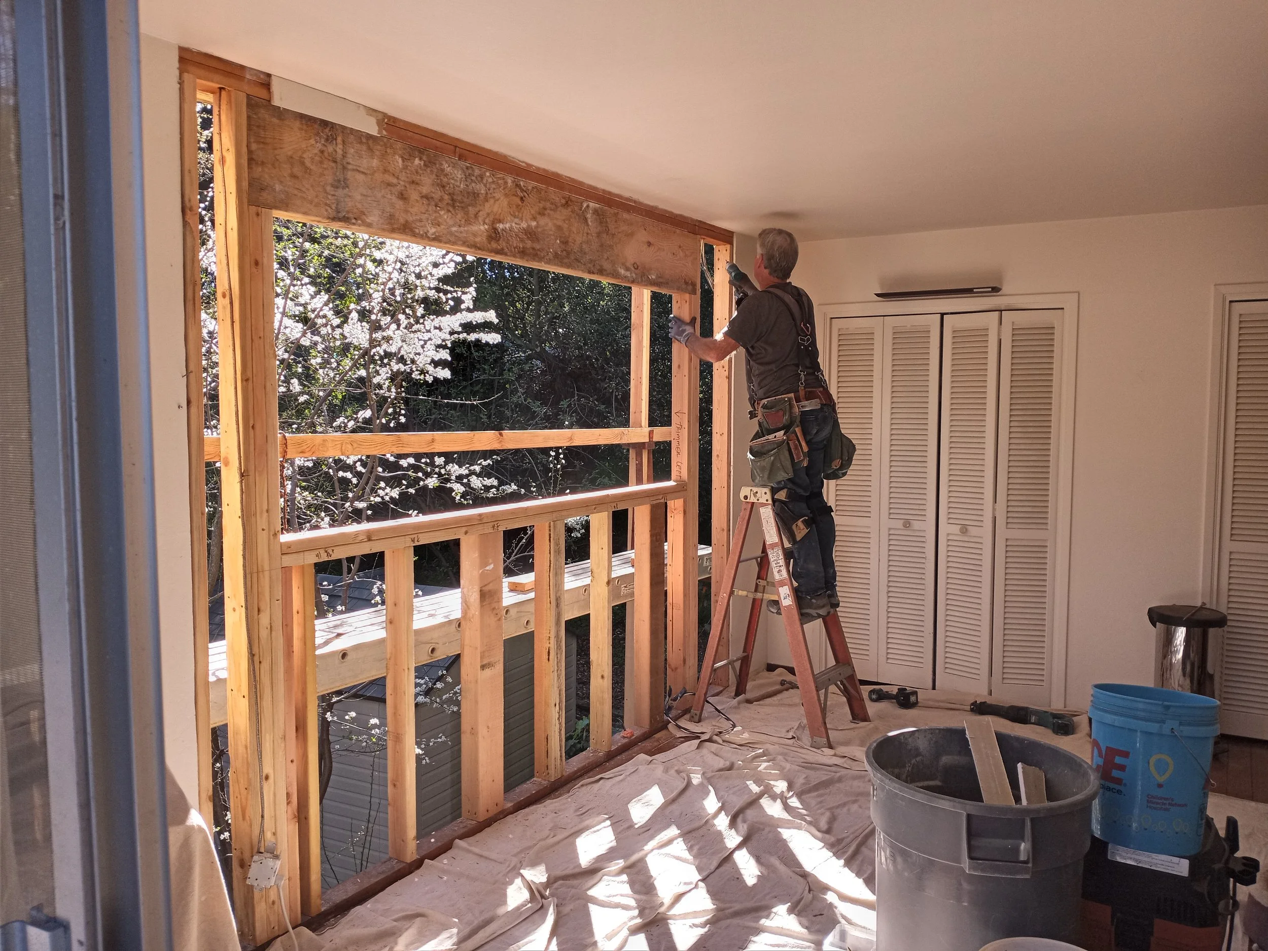 A man on a ladder installing a large window frame on a covered outdoor porch area, with construction tools and materials nearby.