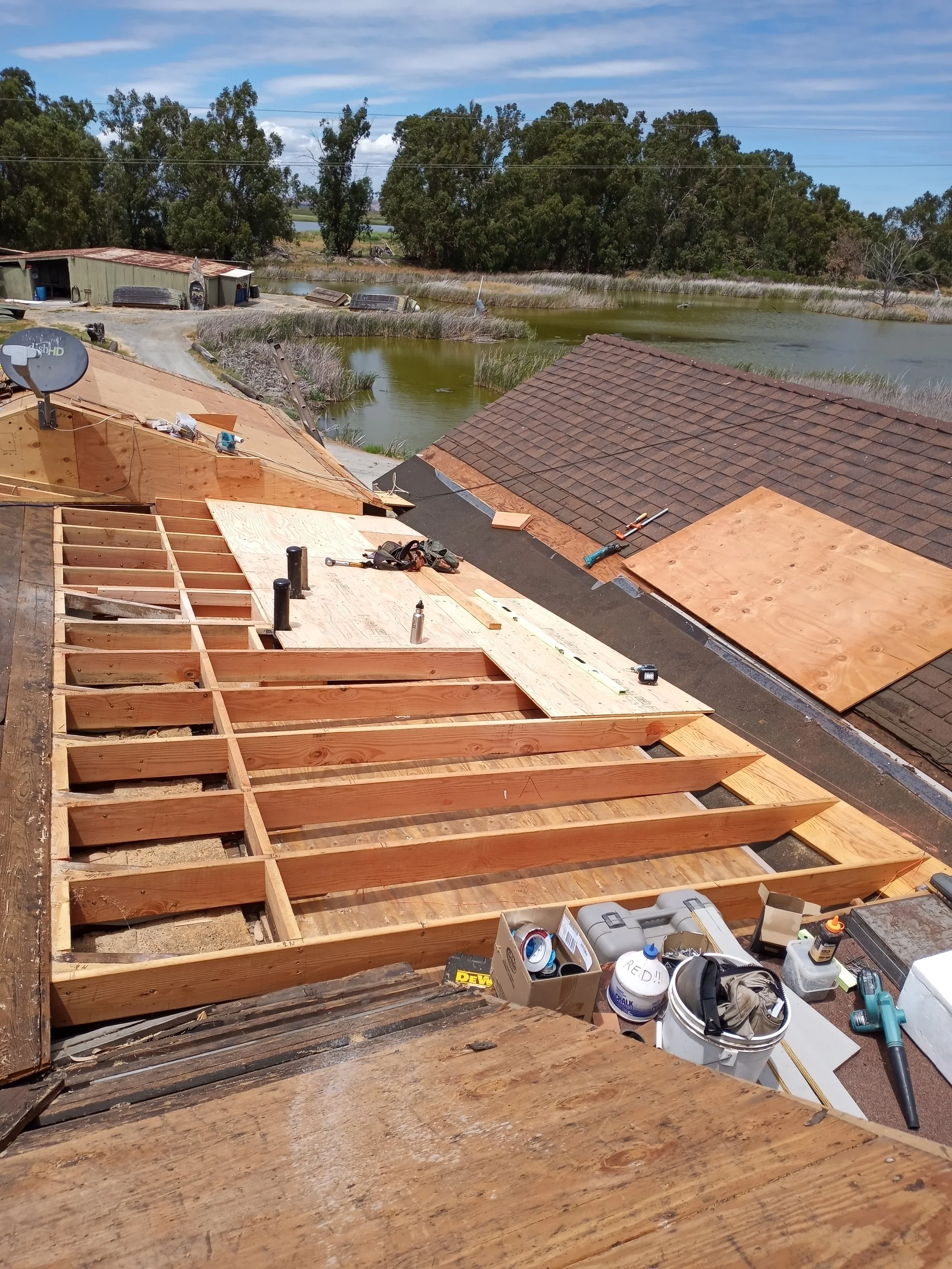 Construction site showing an unfinished wooden deck or roof structure with tools and materials, overlooking lakes and trees under a partly cloudy sky.