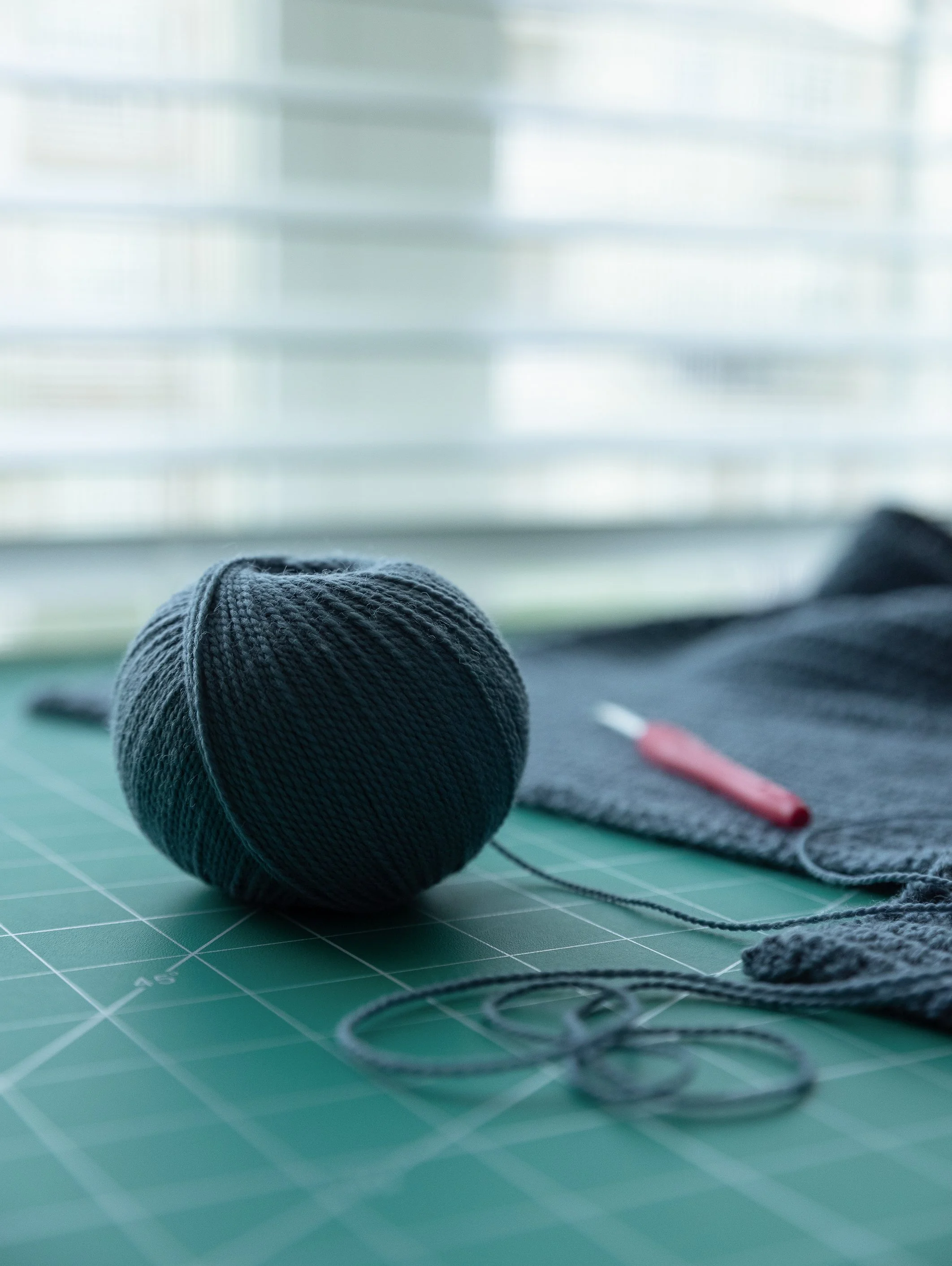 A black knitted hat and a gray fabric mask with a red tool on an orange cutting mat with a green grid, in a craft or sewing workspace.