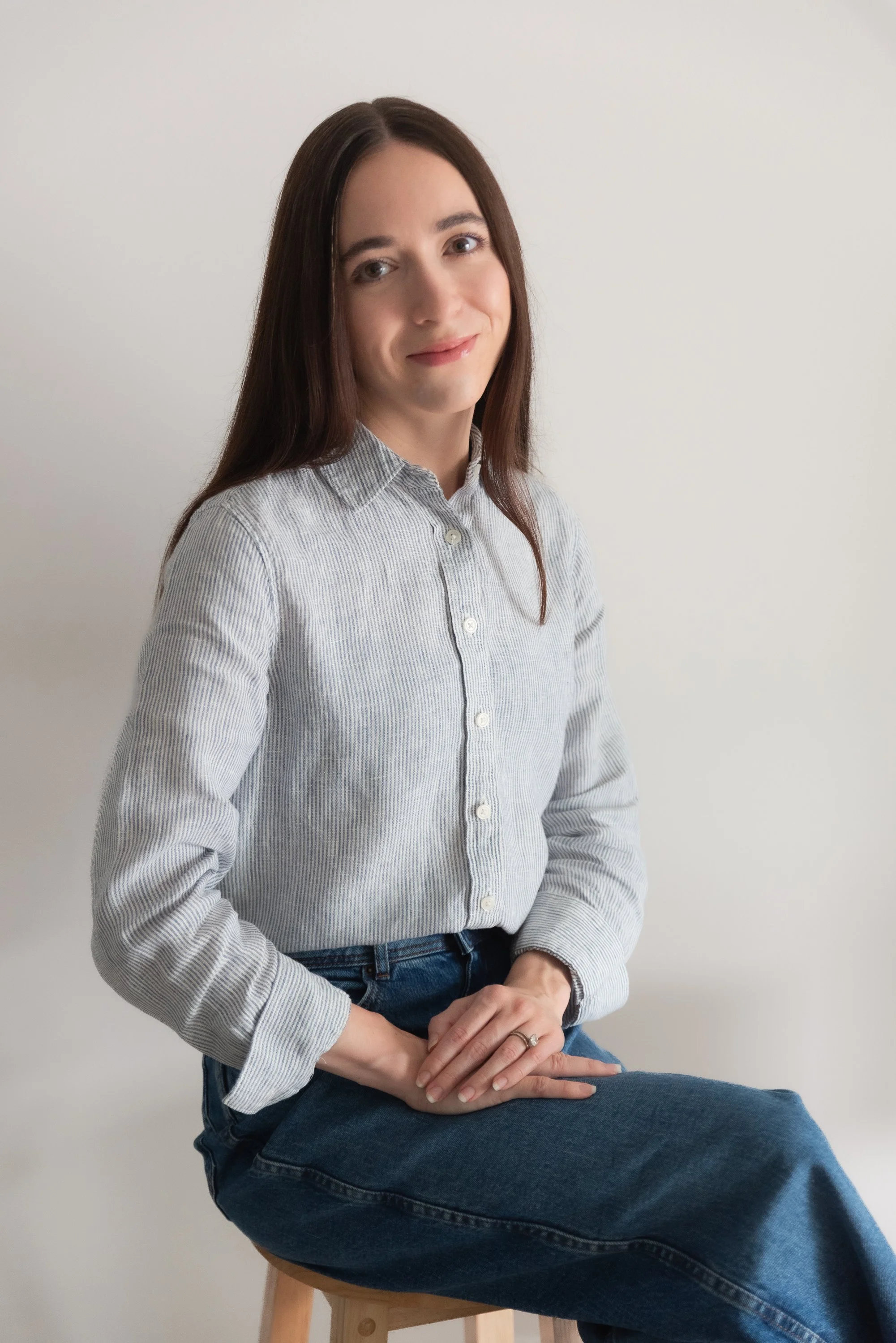 A woman with long brown hair, wearing a light gray striped shirt and blue jeans, seated on a wooden stool with a white background.