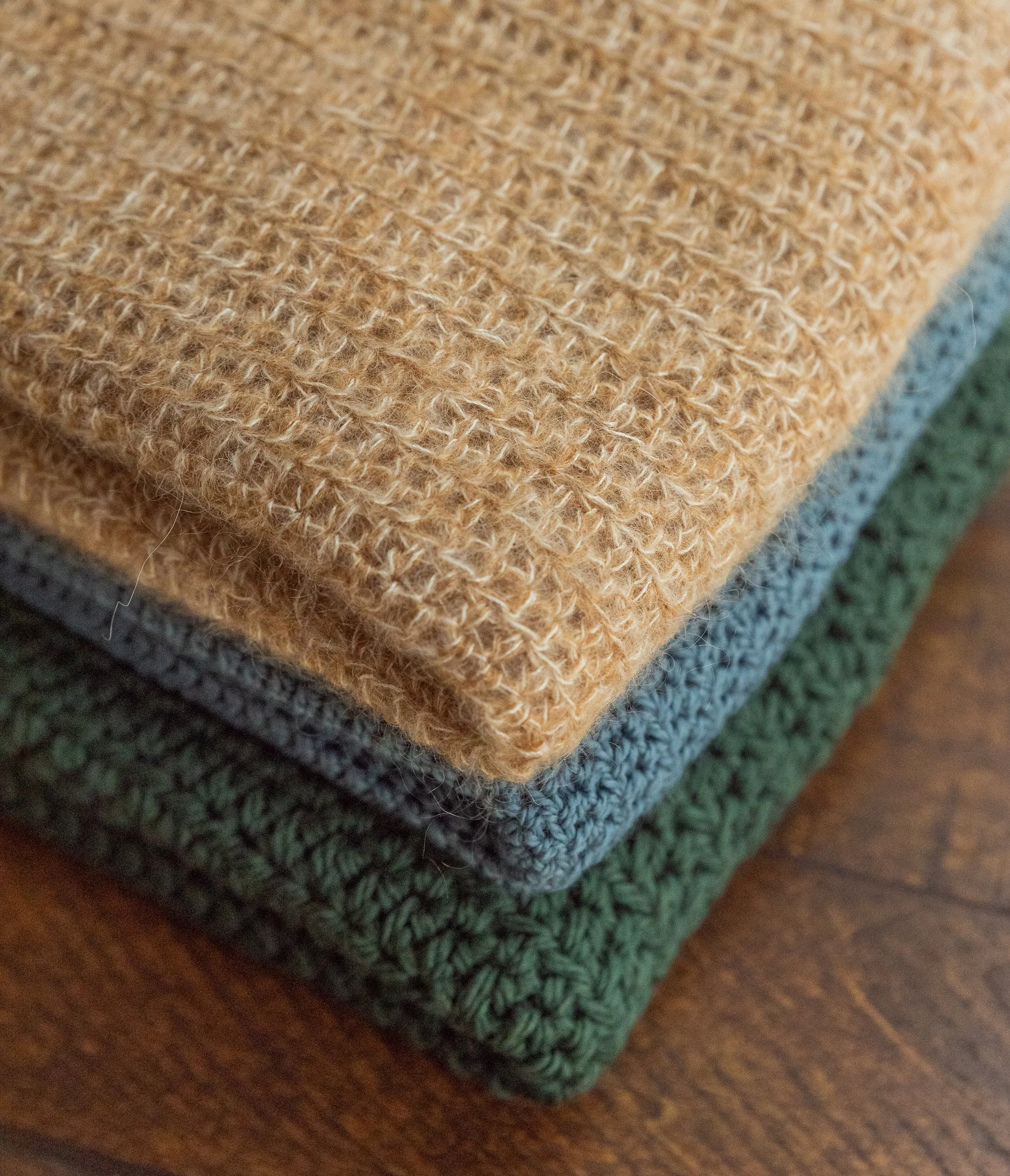Close-up of three folded crochet vests in shades of beige, blue, and dark green on a wooden surface.