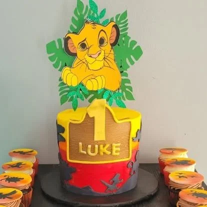 Birthday cake with lion cub theme, decorated with green leaves and a large lion cub topper, with cupcakes around it featuring similar lion designs.