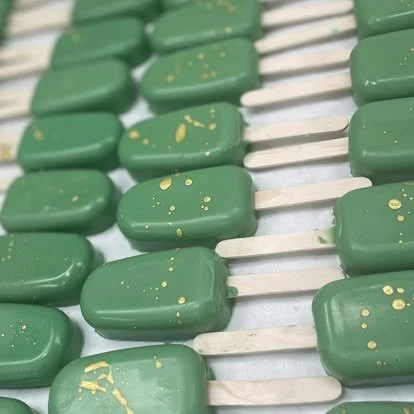 Close-up of green popsicles with gold speckles on white sticks, lined up on a surface.