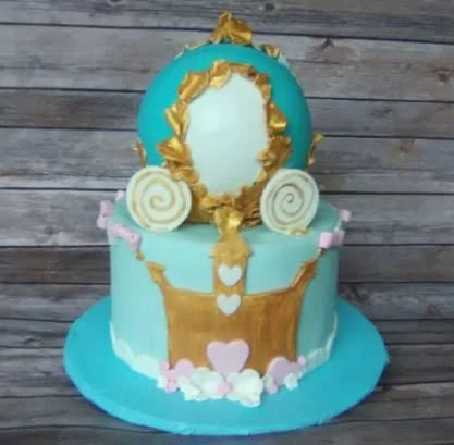 A two-tiered cake with a Cinderella theme featuring a large mirror on top, gold and pink decorations, and white hearts, displayed on a blue cake board against a wooden background.