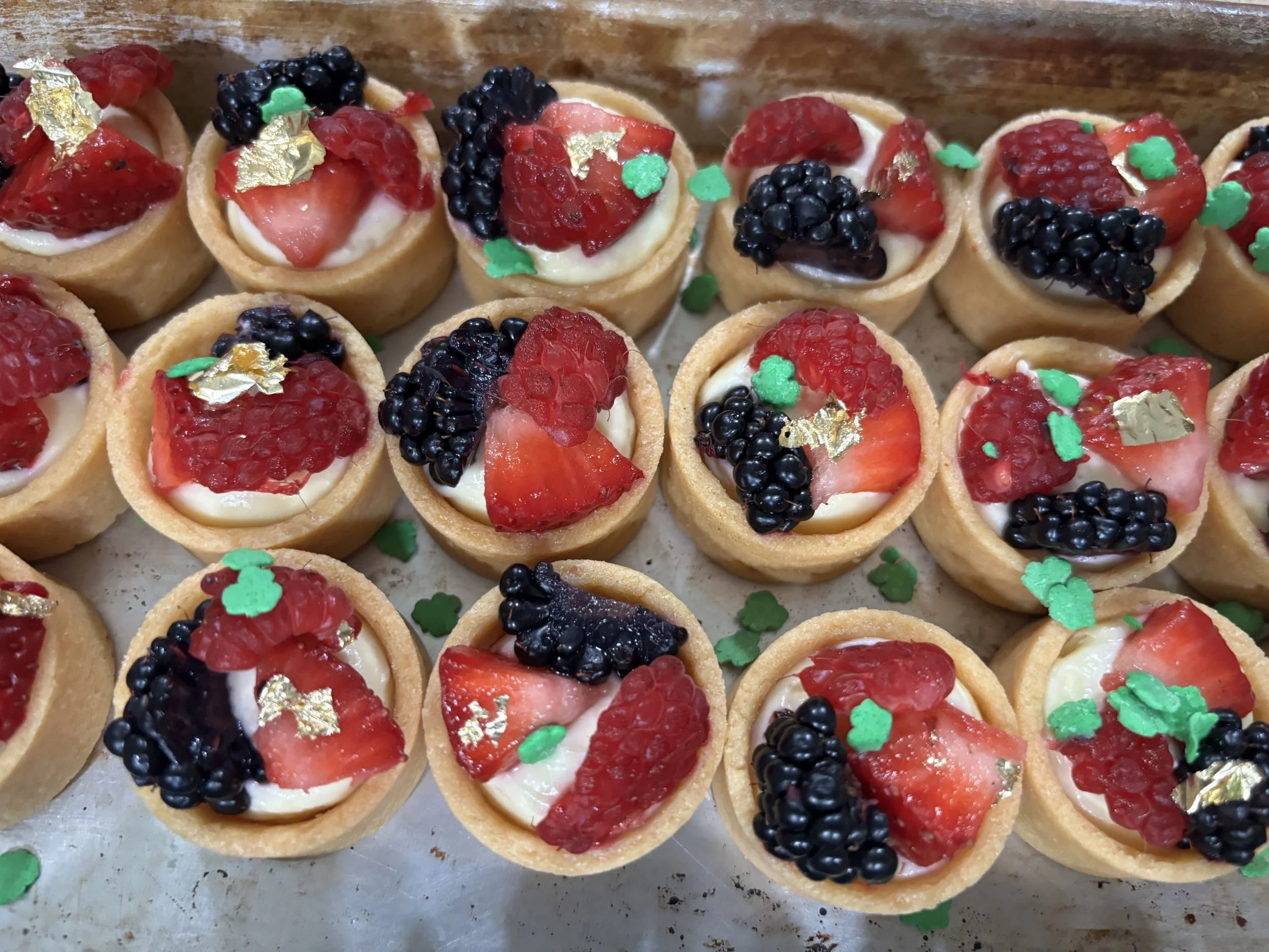 Mini tartlets topped with strawberries, blackberries, red fruit preserves, and gold leaf decorations, with small green shamrock-shaped sprinkles.
