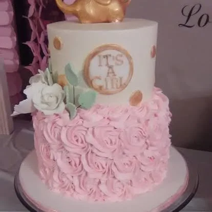 A decorated cake with pink rosette frosting on the bottom and white icing on the top, featuring a gold pig figurine on top and a sign that reads 'It's a Girl'.