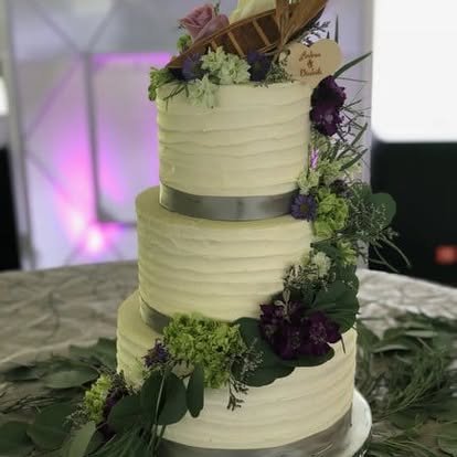 Three-tiered wedding cake decorated with purple and green flowers and foliage, with a black ribbon around each tier, placed on a table with greenery.