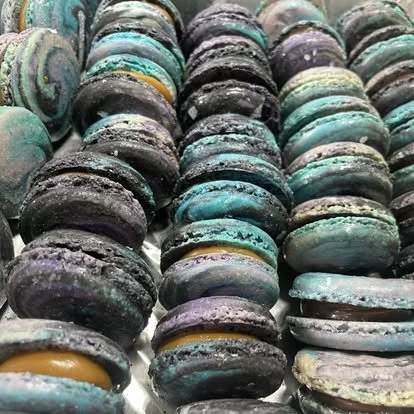 Close-up of colorful macarons in black, purple, turquoise, and multicolored shades, arranged in rows.