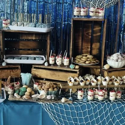 Dessert table with cupcakes, cookies, and parfait glasses, decorated with blue and silver party decorations and a blue tablecloth.