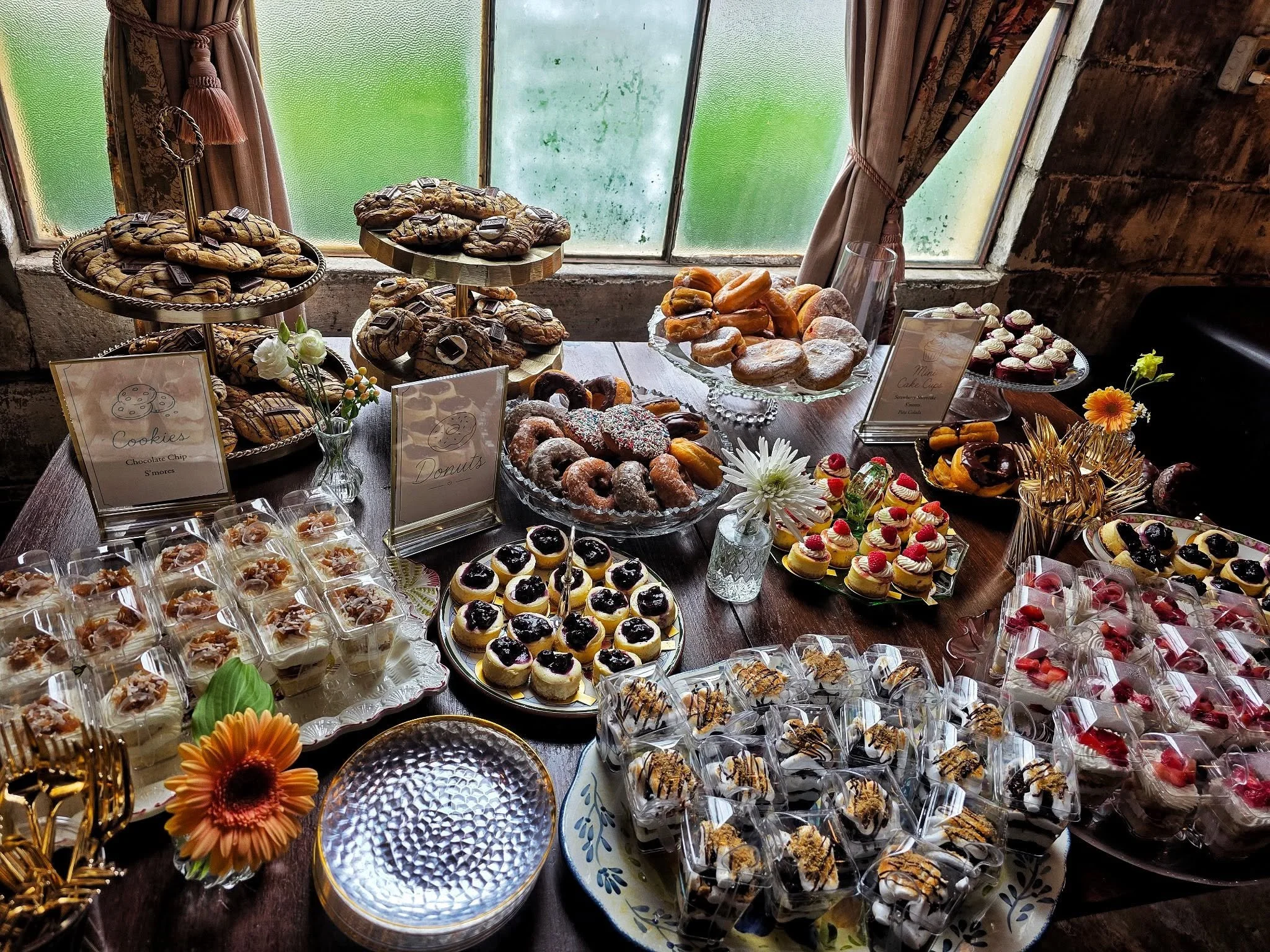 A variety of baked goods and desserts displayed on a table, including cookies, donuts, cupcakes, and other pastries, with decorative flowers and signs labeling some items.