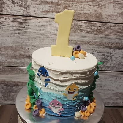 A birthday cake with ocean-themed decorations, a large number 1 candle on top, and colorful fondant sharks and seaweed around the sides.
