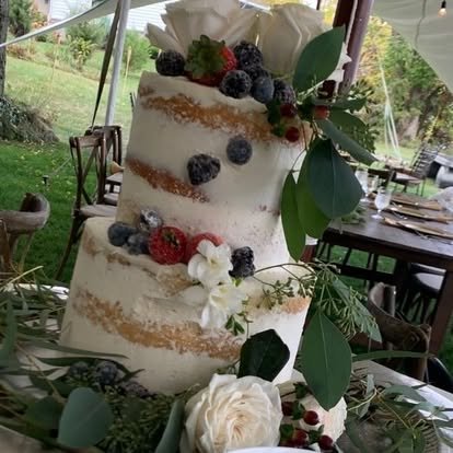 A tall, tiered naked cake decorated with fresh berries and white flowers, set outdoors on a table with greenery.
