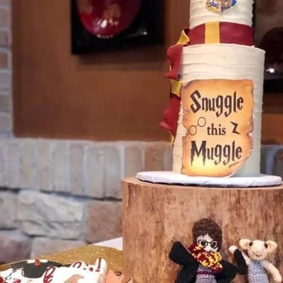 A cake with a Harry Potter-themed topper featuring a sign that says 'Snuggle this Muggle,' and two stuffed animals in front of it, placed on a Wooden cake stand.