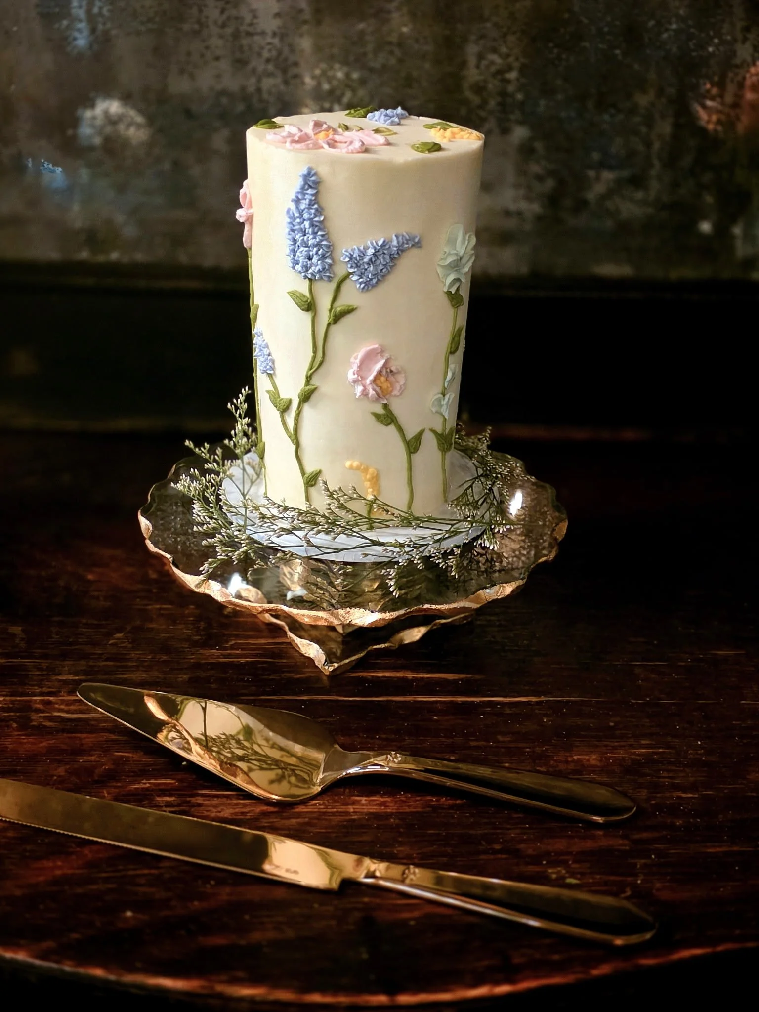 A tall white cake decorated with pastel-colored flowers and green leaves, sitting on a silver platter, with a gold-colored cake knife and server in front of it on a dark wood table.
