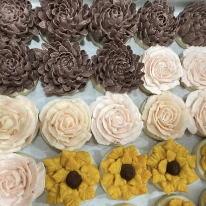 Assorted flower-shaped and sunflower-shaped cupcakes arranged in rows.
