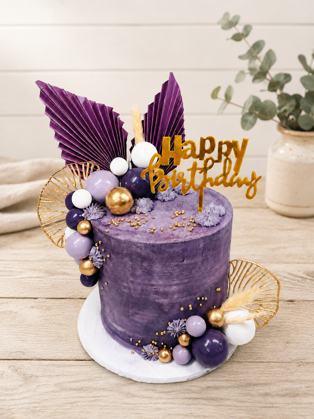Purple birthday cake decorated with gold, white, and purple spherical ornaments, purple leaves, gold accents, and a gold "Happy Birthday" topper on a wooden table with a plant in a pot in the background.