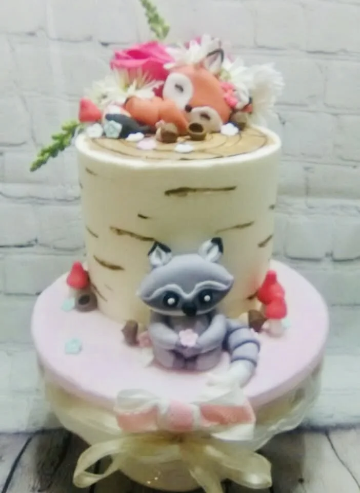 Two-tiered cake decorated with a raccoon figure on the bottom tier and a fox on the top, with flowers, small figurines, and decorative elements, against a white brick wall background.