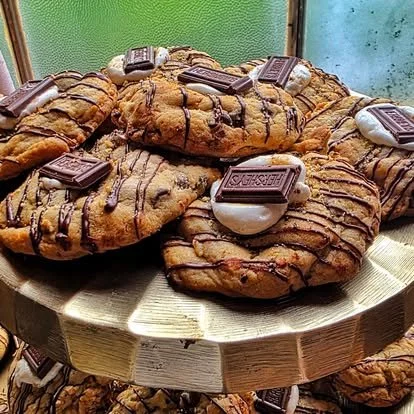 A pile of large chocolate chip cookies topped with melting ice cream, chocolate drizzle, and pieces of Snickers candy bars on a wooden stand.