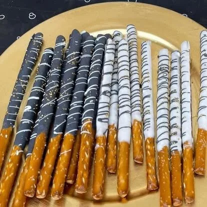 A golden tray with chocolate-dipped pretzel sticks, some topped with white or dark chocolate drizzle.