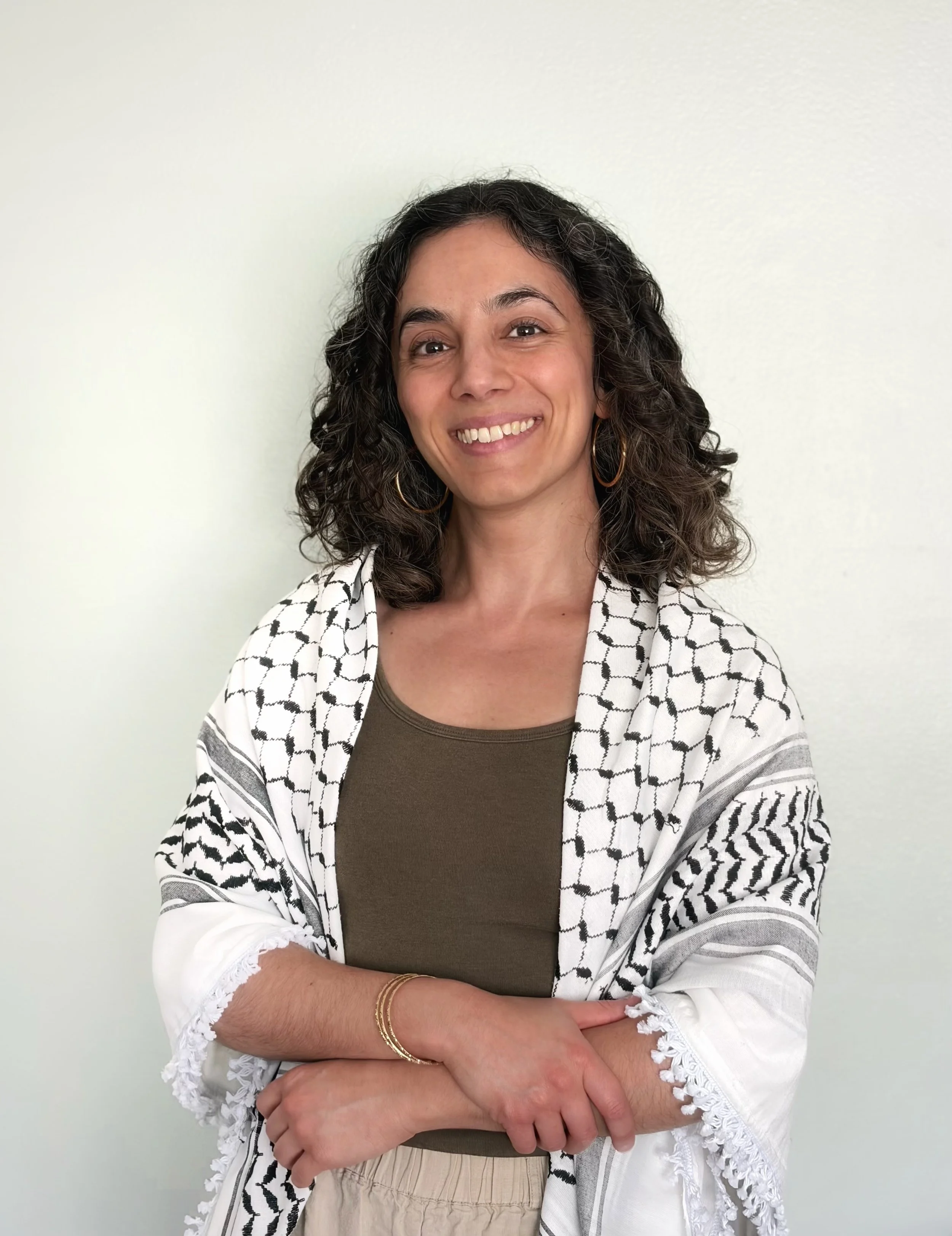 A woman with curly dark hair and hoop earrings smiling, wearing a brown top and beige pants, standing against a plain white wall.