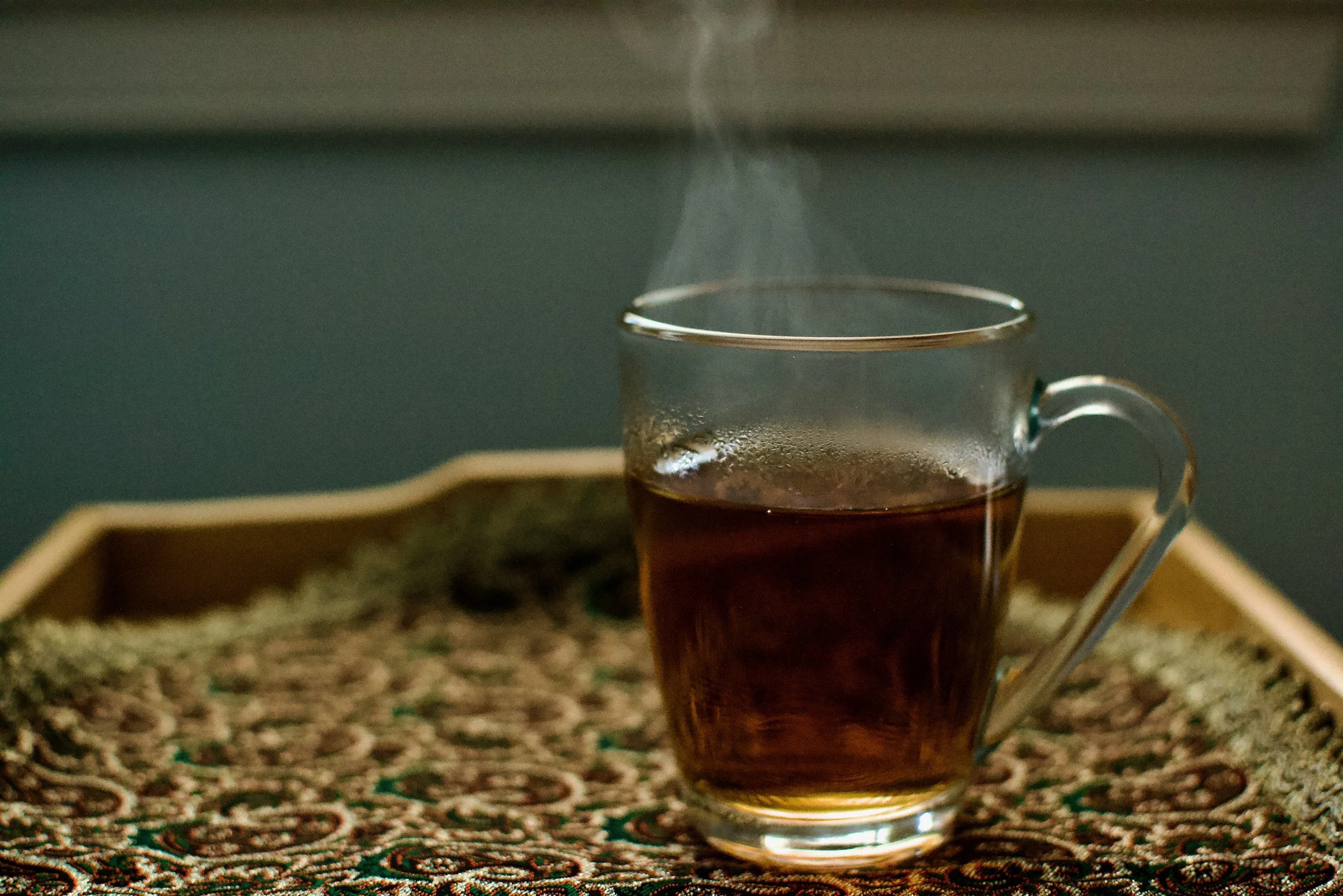 A glass cup filled with a dark beverage, possibly tea or coffee, on a decorative patterned tablecloth.