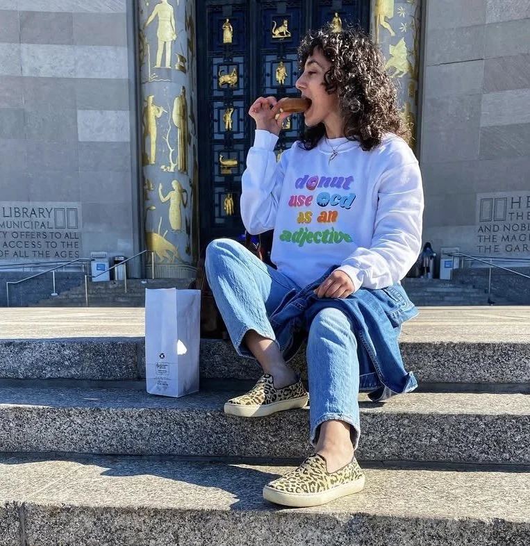 A woman with dark hair sitting on the steps in front of a shop, eating a donut. She is wearing a white sweatshirt that says "donut use OCD as an adjective." There is a white paper bag next to her.