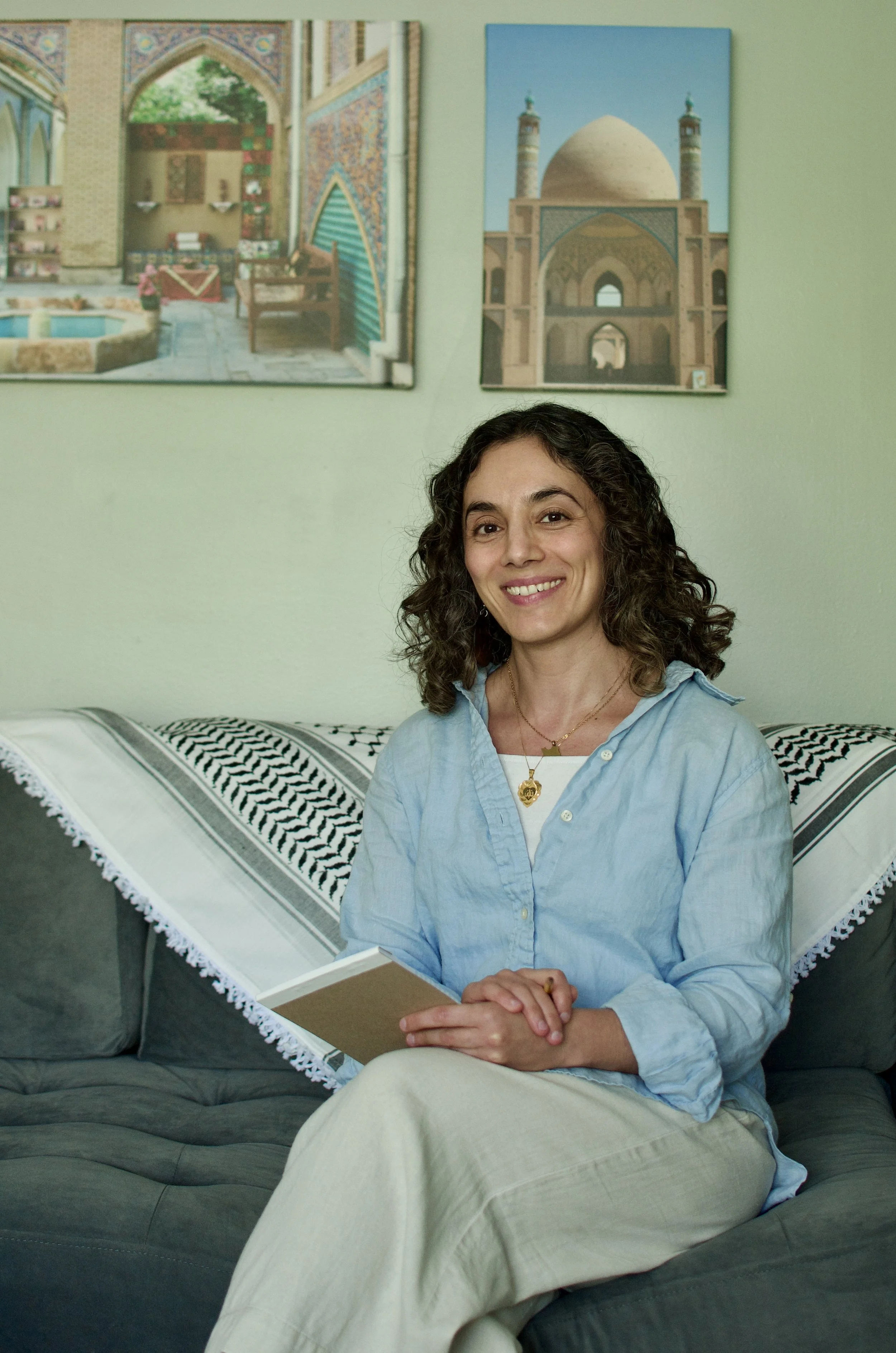 A woman with curly brown hair and light skin sitting on a gray sofa, smiling at the camera, wearing a light blue shirt and holding a closed notebook. Behind her are two framed pictures of architectural landmarks, and a blanket with black and white geometric patterns draped over the sofa.