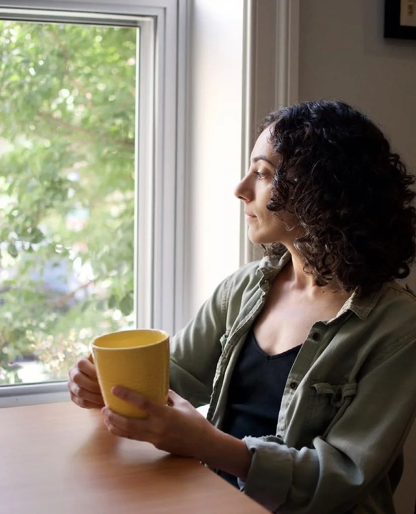 Woman with curly dark hair sitting at a wooden table near a window, holding a yellow mug and looking outside, wearing a dark shirt and a green jacket.