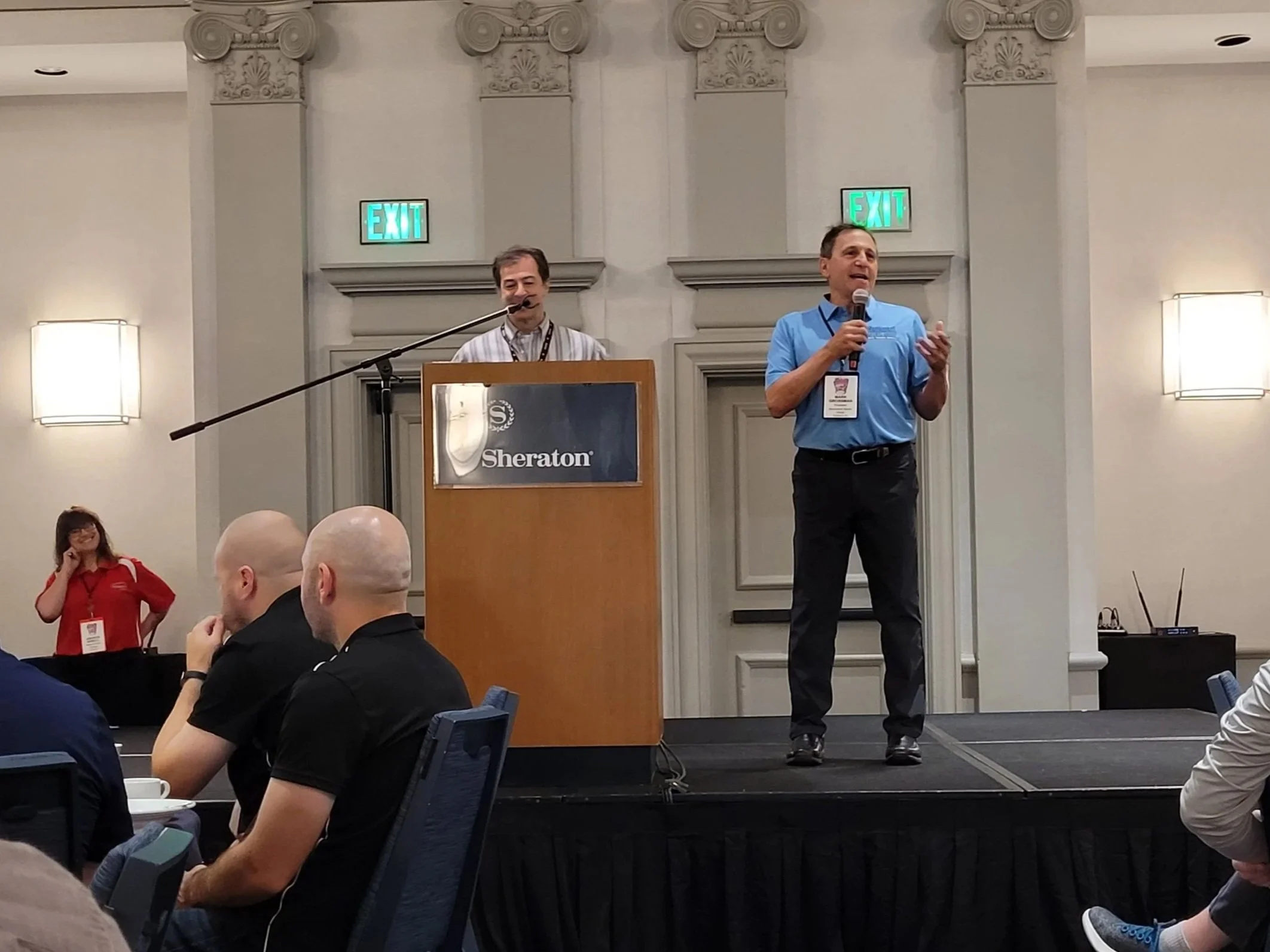 Two men are speaking at a conference. One is standing and speaking into a microphone, wearing a blue shirt, and the other is standing behind a podium, wearing a striped shirt. The background features decorative columns, two exit signs, and conference signage at the Sheraton hotel.