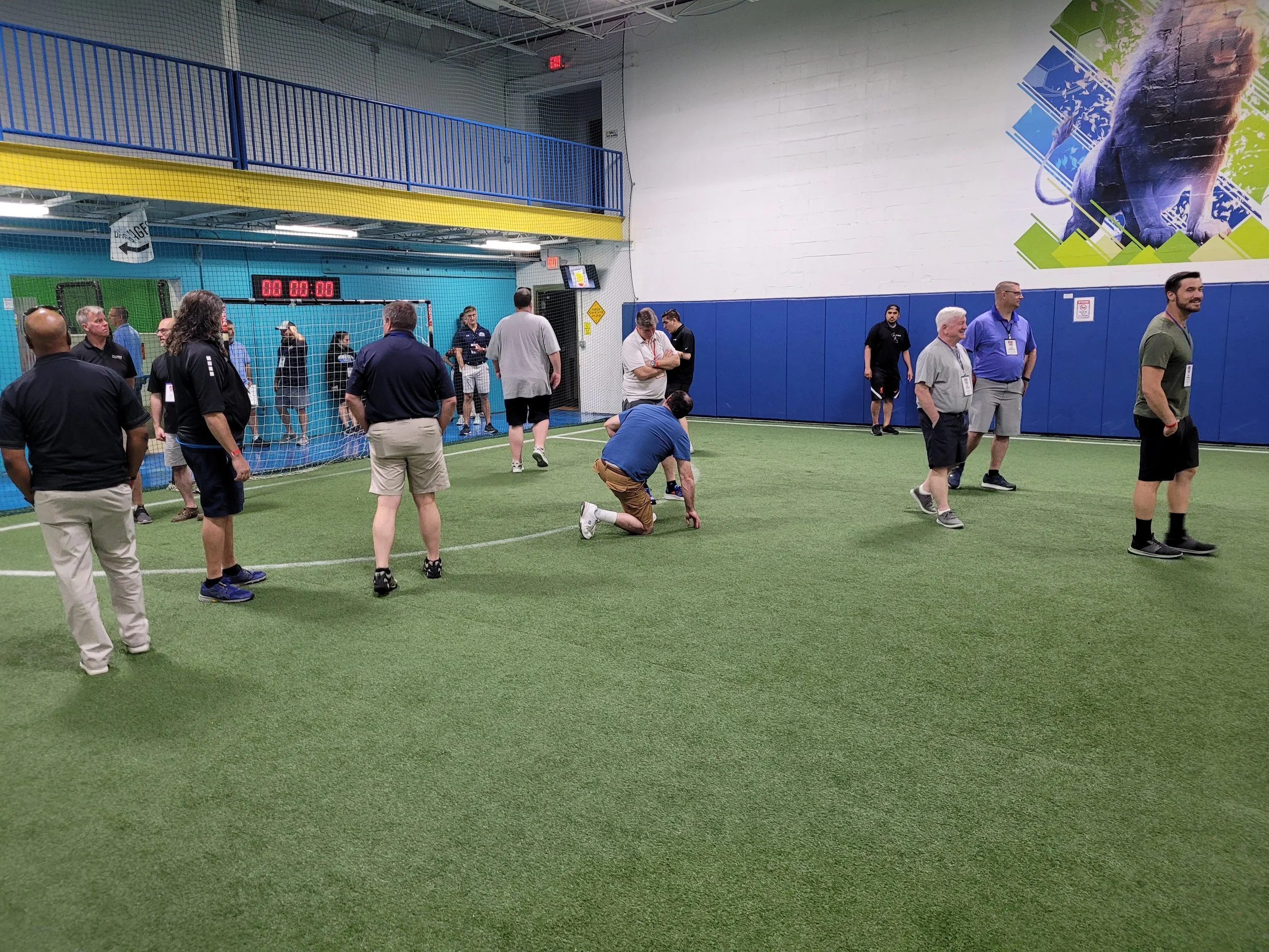 Indoor sports facility with artificial turf, people standing and walking, a netted area, and an electronic scoreboard showing all zeros. There is a painted lion on the wall.