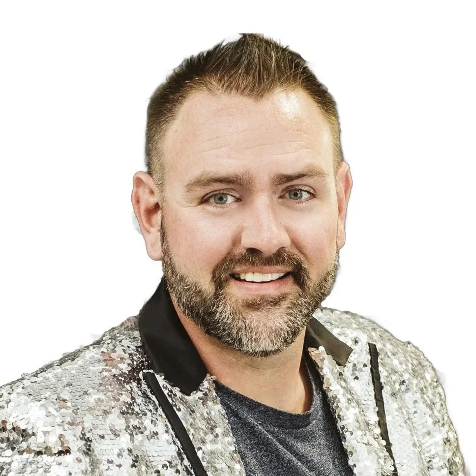 A man with a beard and short hair, smiling, wearing a gray shirt and a shiny, silver sequined jacket.