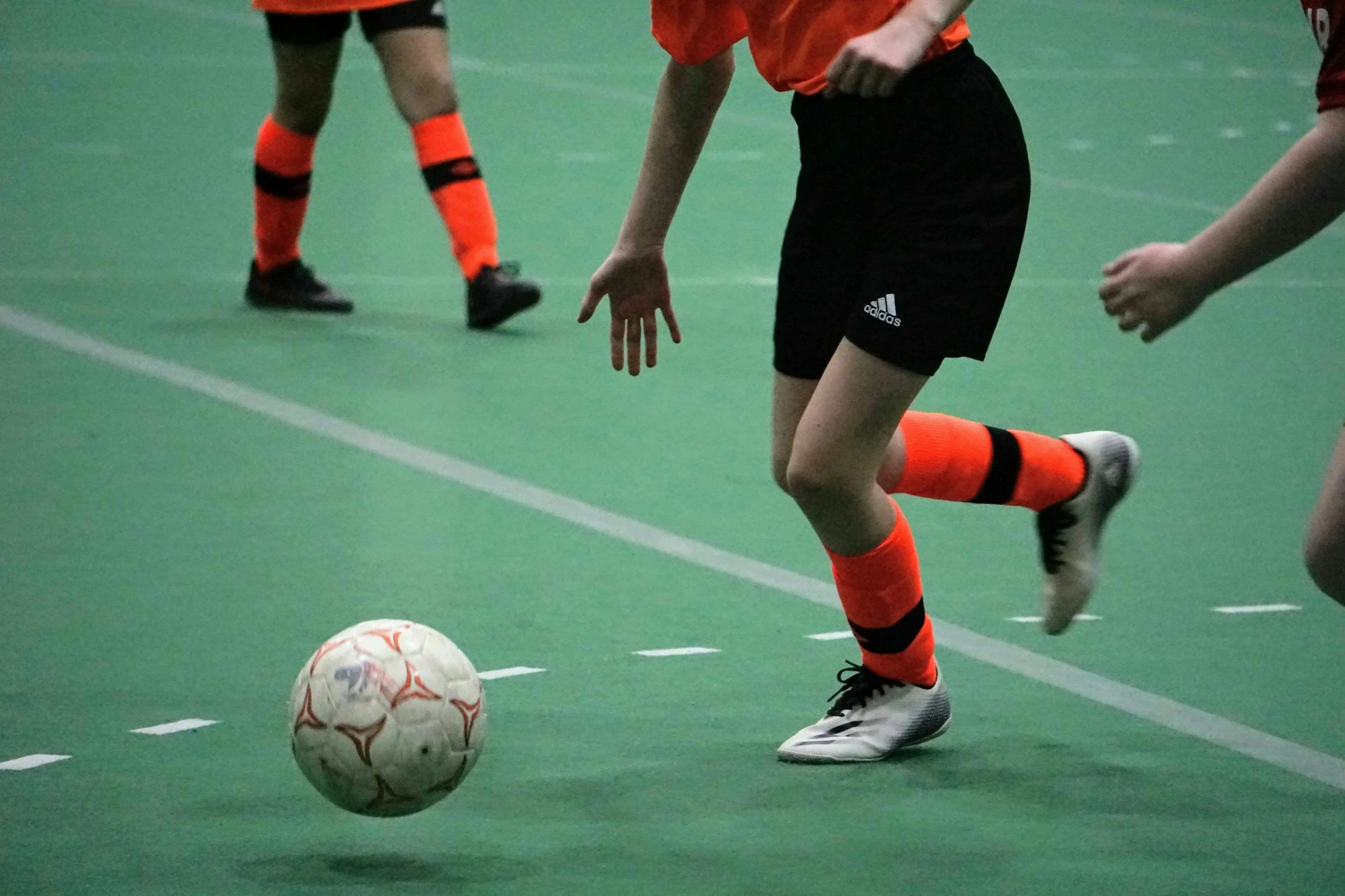 Soccer players on an indoor field wearing black and orange kits, with one player about to kick a ball.