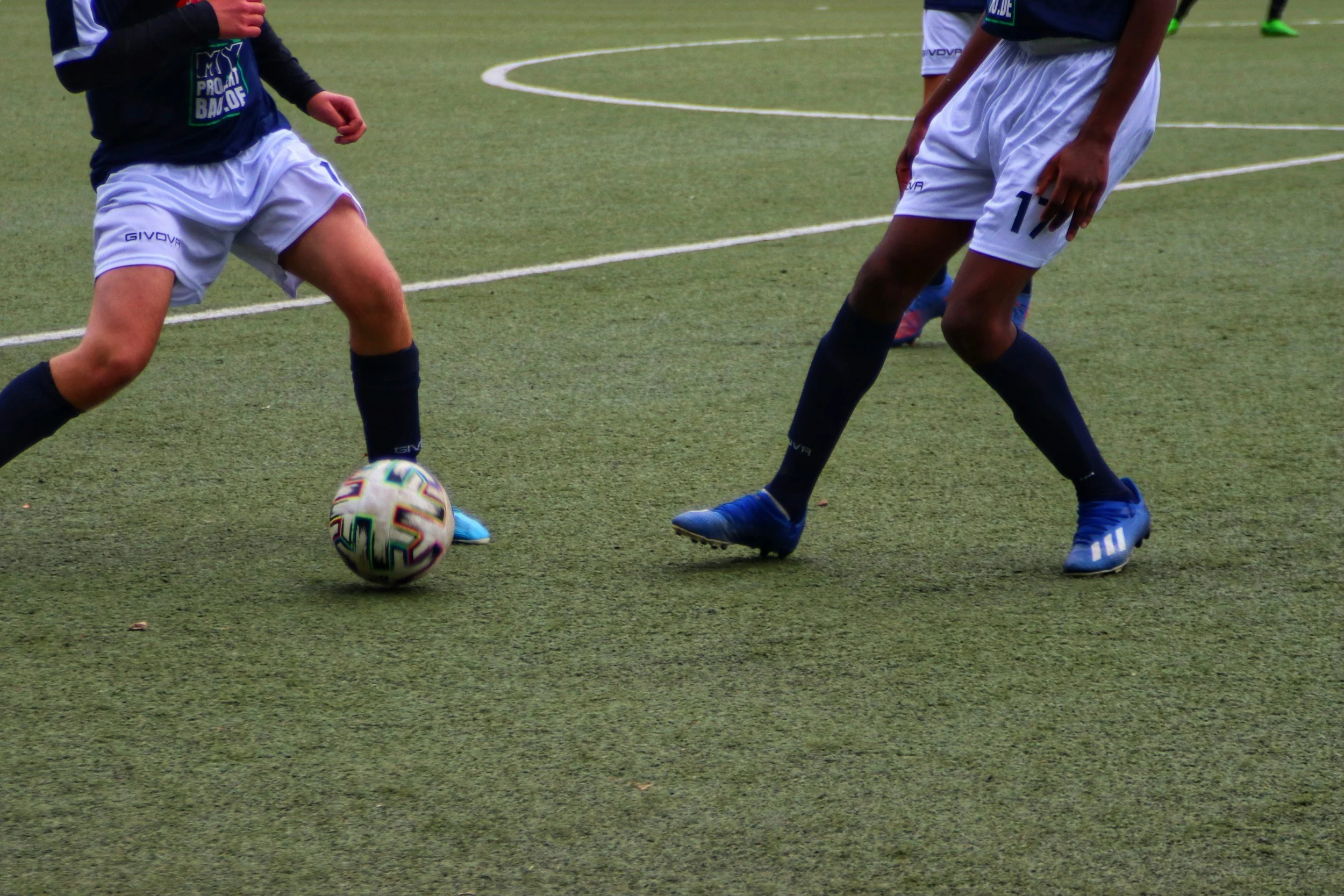 Two soccer players are competing for the ball on a green turf field. One player is dressed in a dark shirt and white shorts, and another in a blue shirt and white shorts. The soccer ball is close to the feet of the player in the dark shirt.