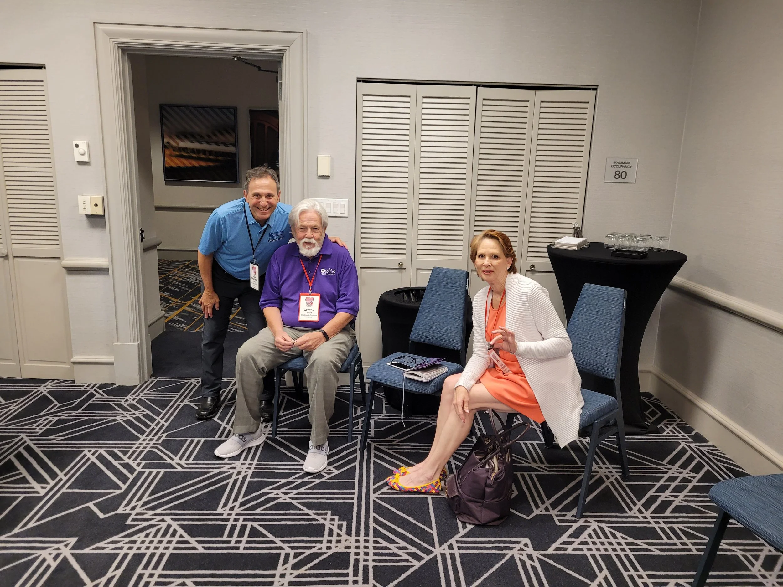 Three people at a conference or event in a hotel ballroom, two men and one woman, posing for a photo. The man on the left is wearing a blue shirt and black pants, smiling and standing. The man in the middle is seated, has white hair and a beard, wear