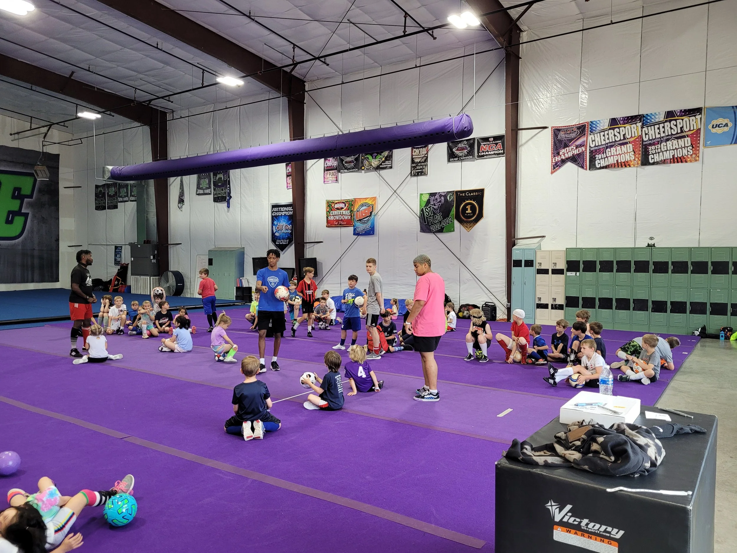Kids and coaches sitting and standing on a purple mat inside a gym during a sports practice or camp, with banners and lockers on the wall.