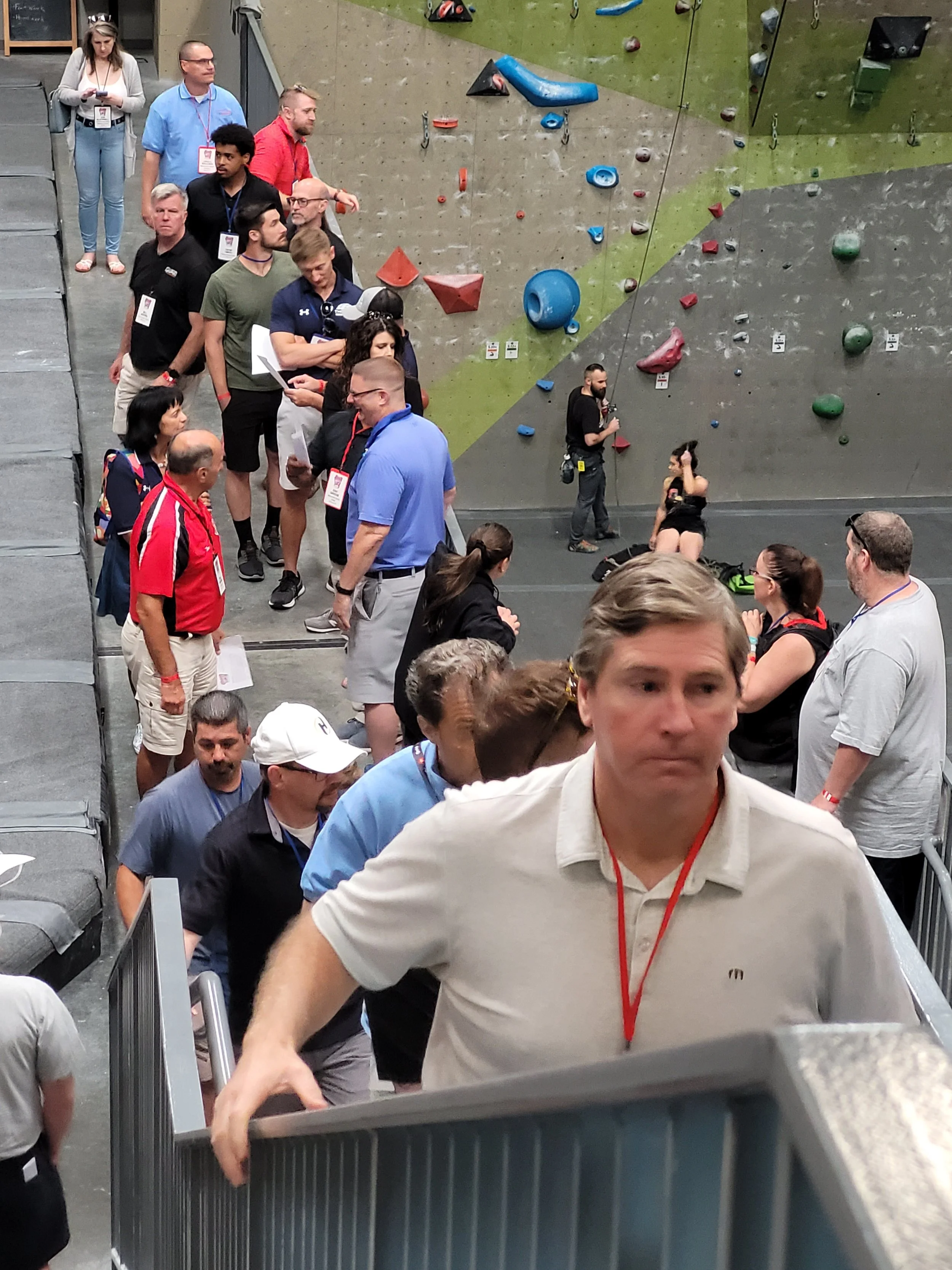 A group of people waiting in line at an indoor climbing gym near the climbing wall. Some are engaged in conversation and others are focused on their activities.