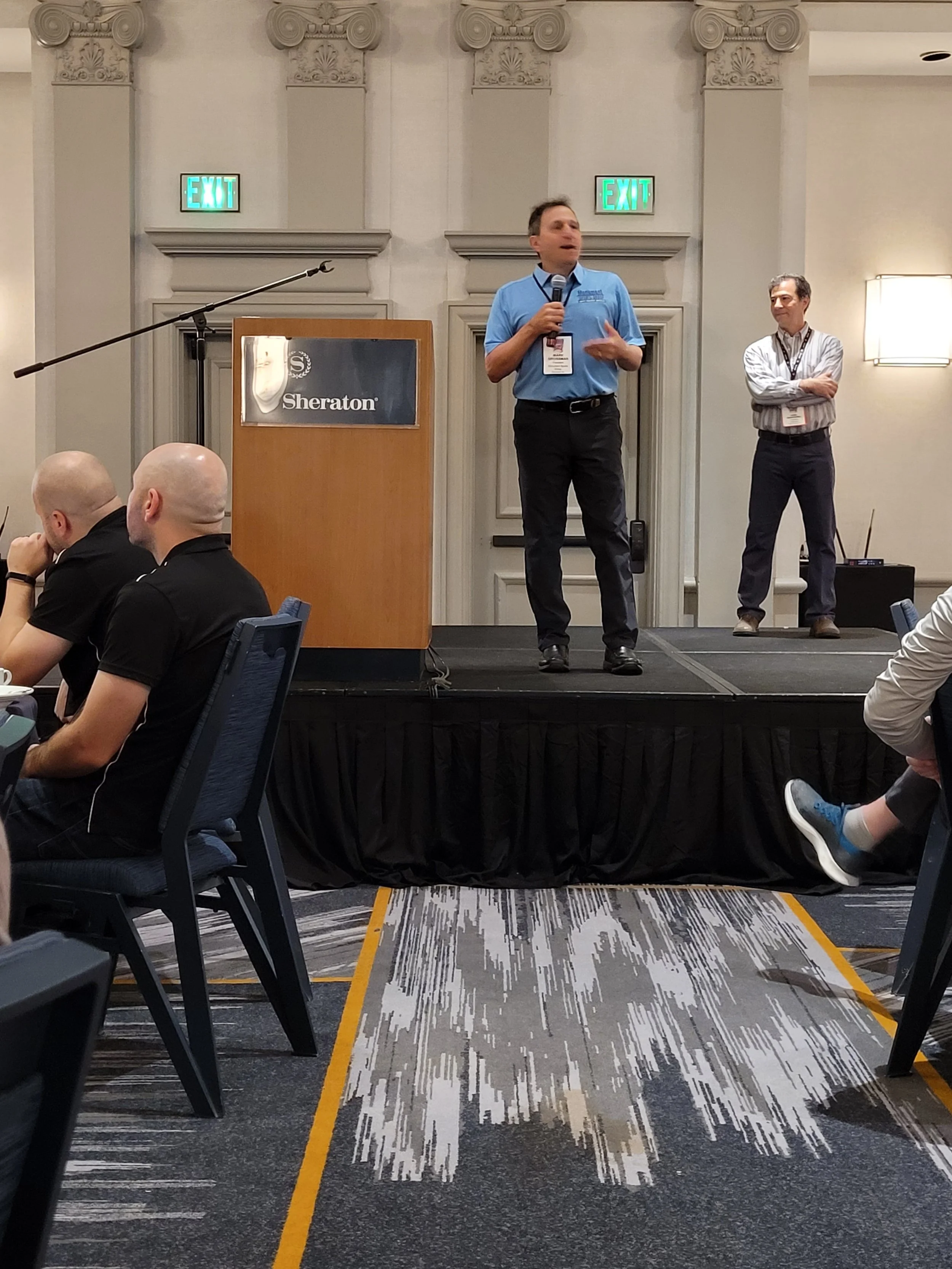 A man stands on a stage holding a microphone and speaking at a conference in a hotel conference room. There is a wooden podium with a Sheraton logo to his left. Behind him, another man stands with his arms crossed, listening. The conference room has 