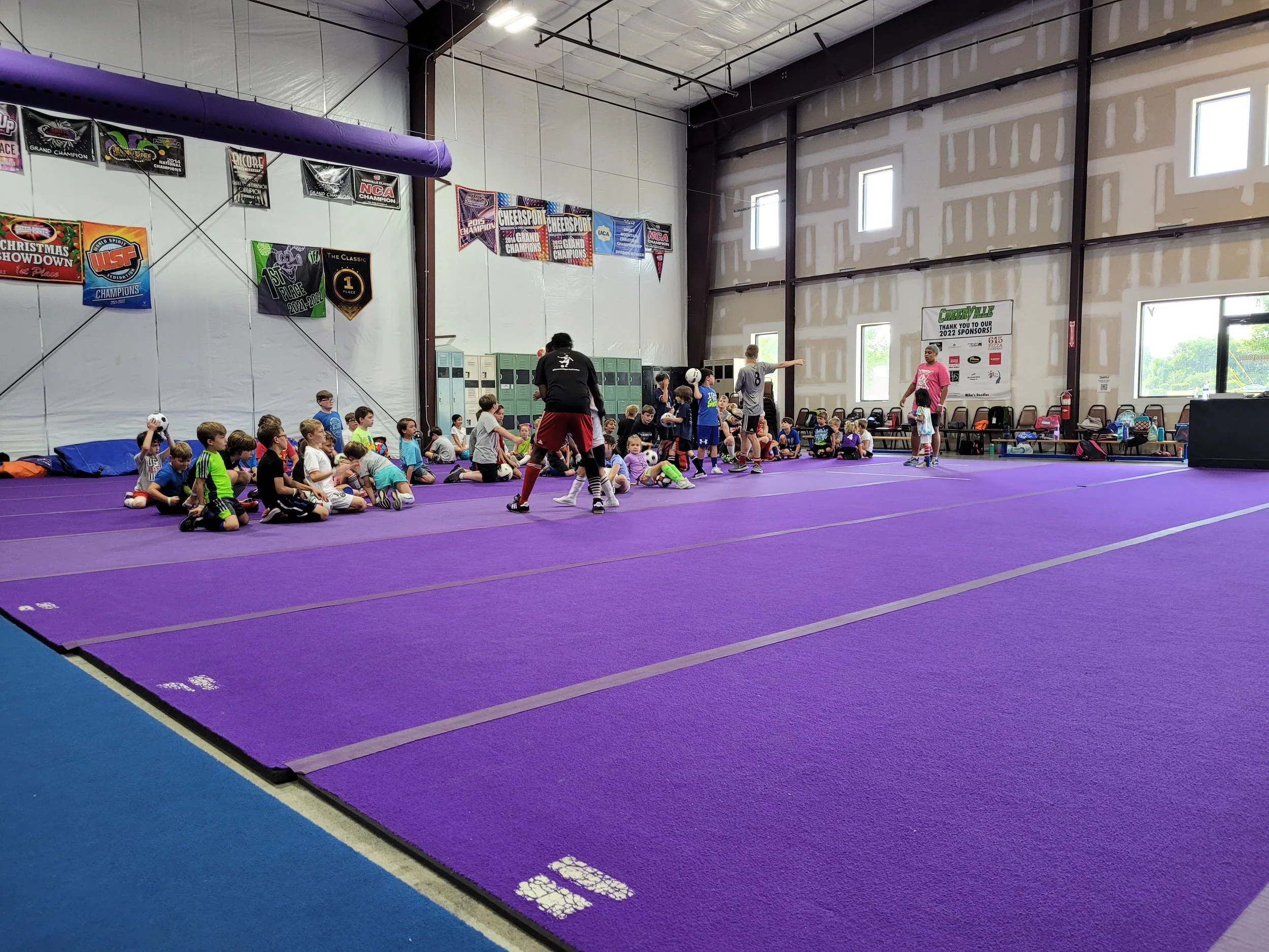 Children sitting on purple gym mats inside a gymnasium, with some adults supervising, engaged in sports activities. Banners and flags hang on the walls.