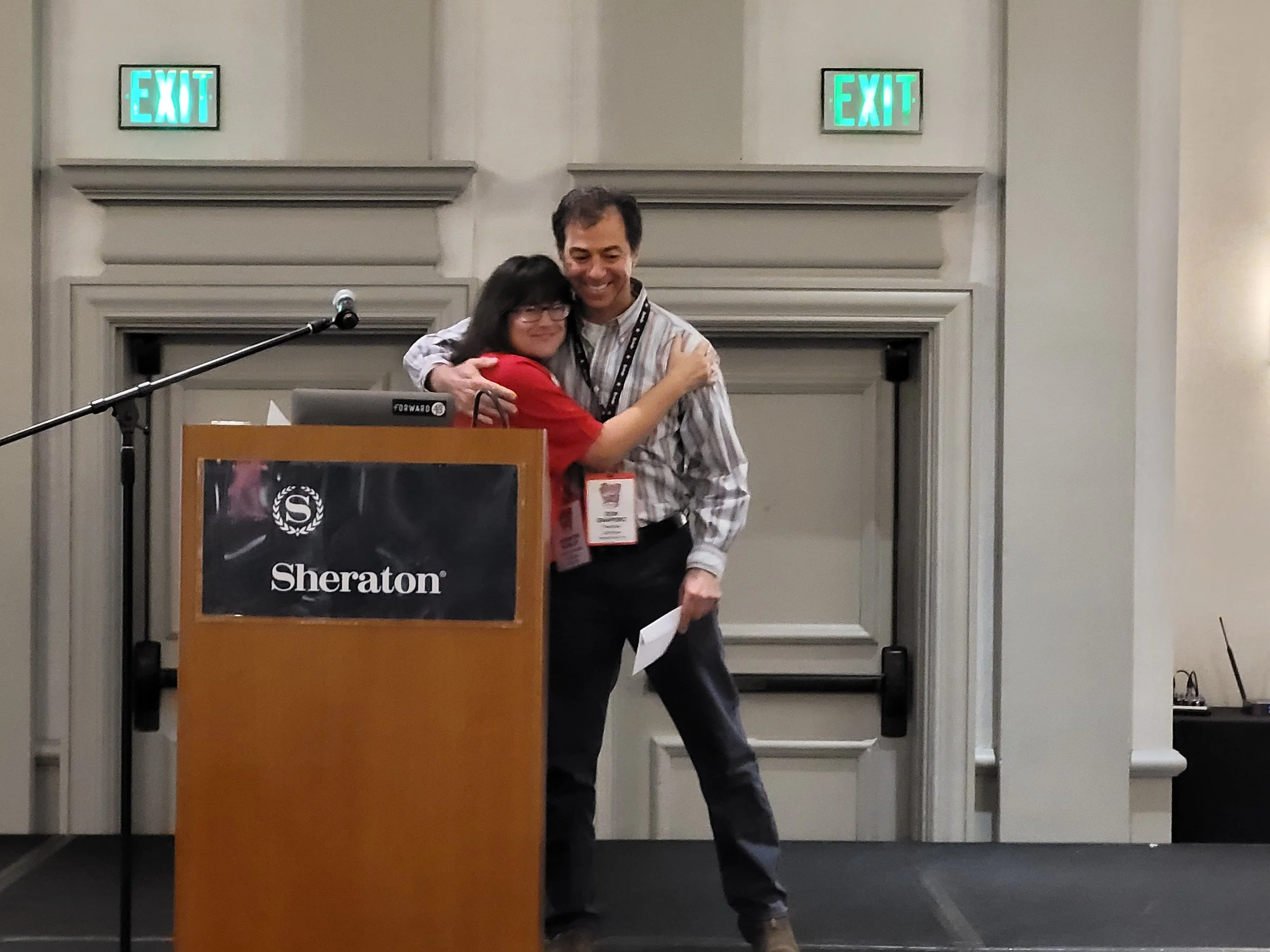 Two people are hugging in front of a podium at a conference in a hotel ballroom. There are two illuminated exit signs on the wall behind them.