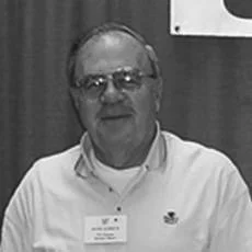 An older man with glasses and a mustache, wearing a white collared shirt, sitting in front of a wooden background.