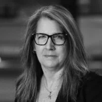 A woman with shoulder-length hair, glasses, and a serious expression, wearing a dark jacket.